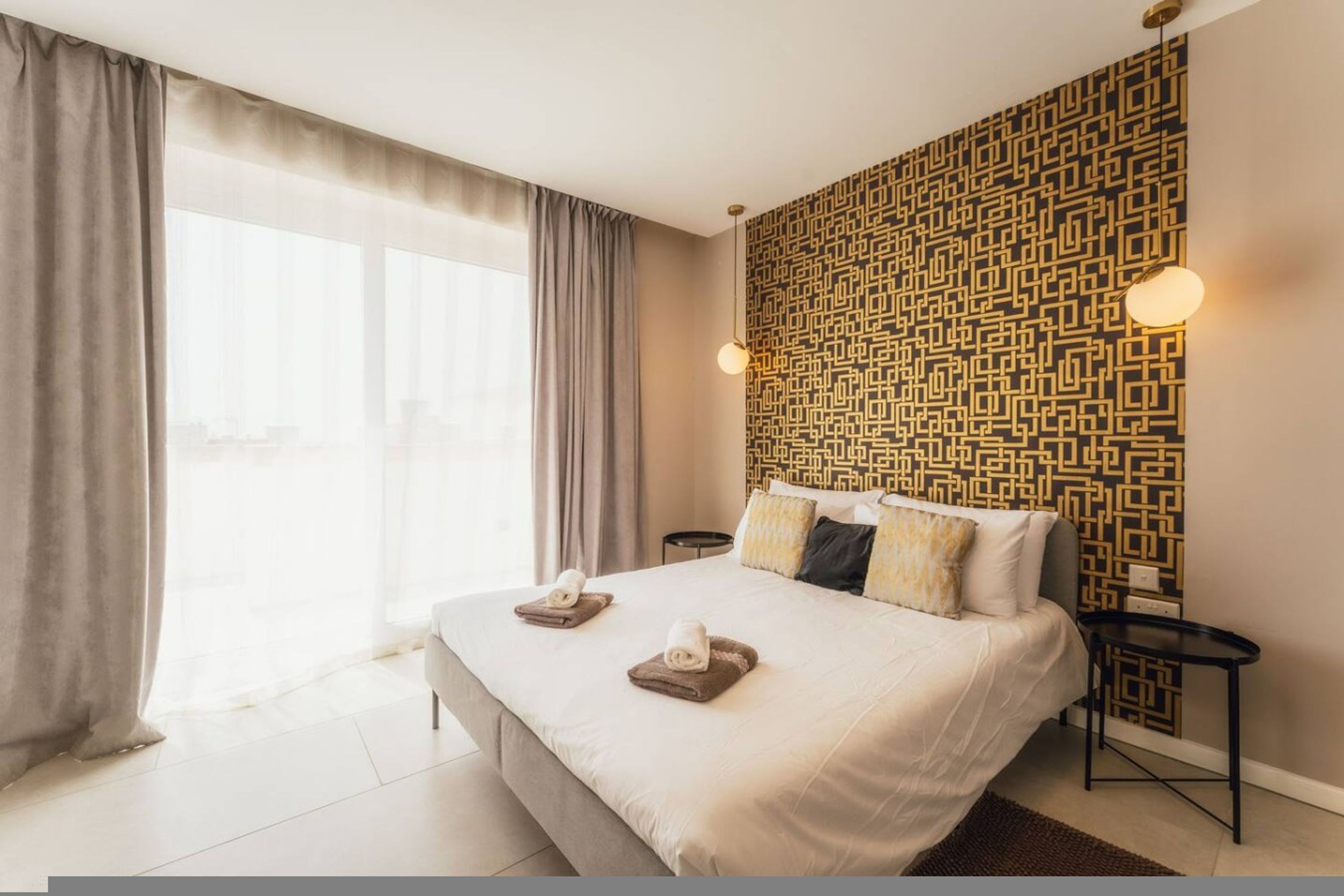 A hotel room with a large bed, two bedside tables, a patterned wall, and a view through the window.