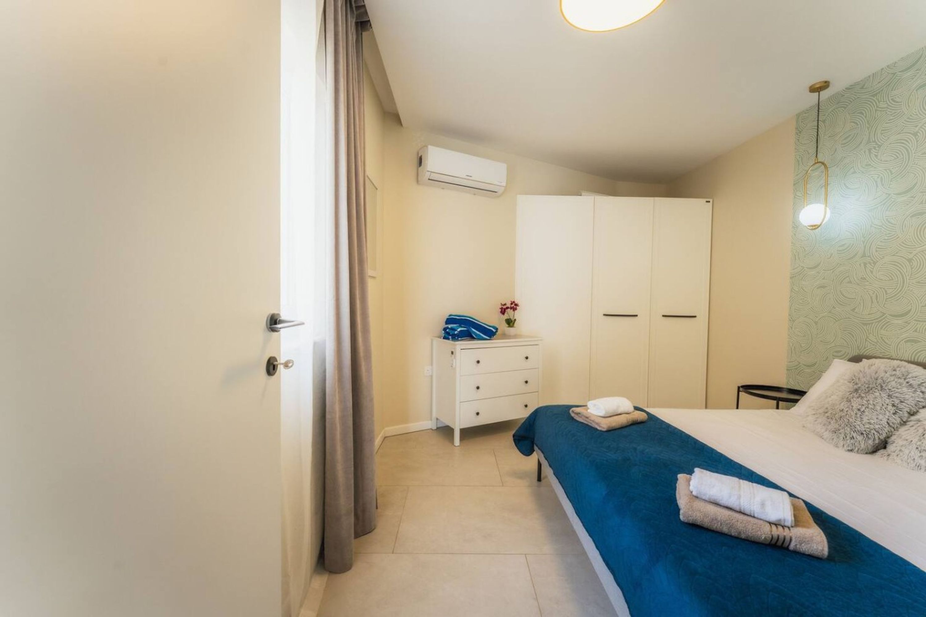 A bedroom with a bed, a dresser, a wardrobe, a nightstand, and an air conditioning unit.
