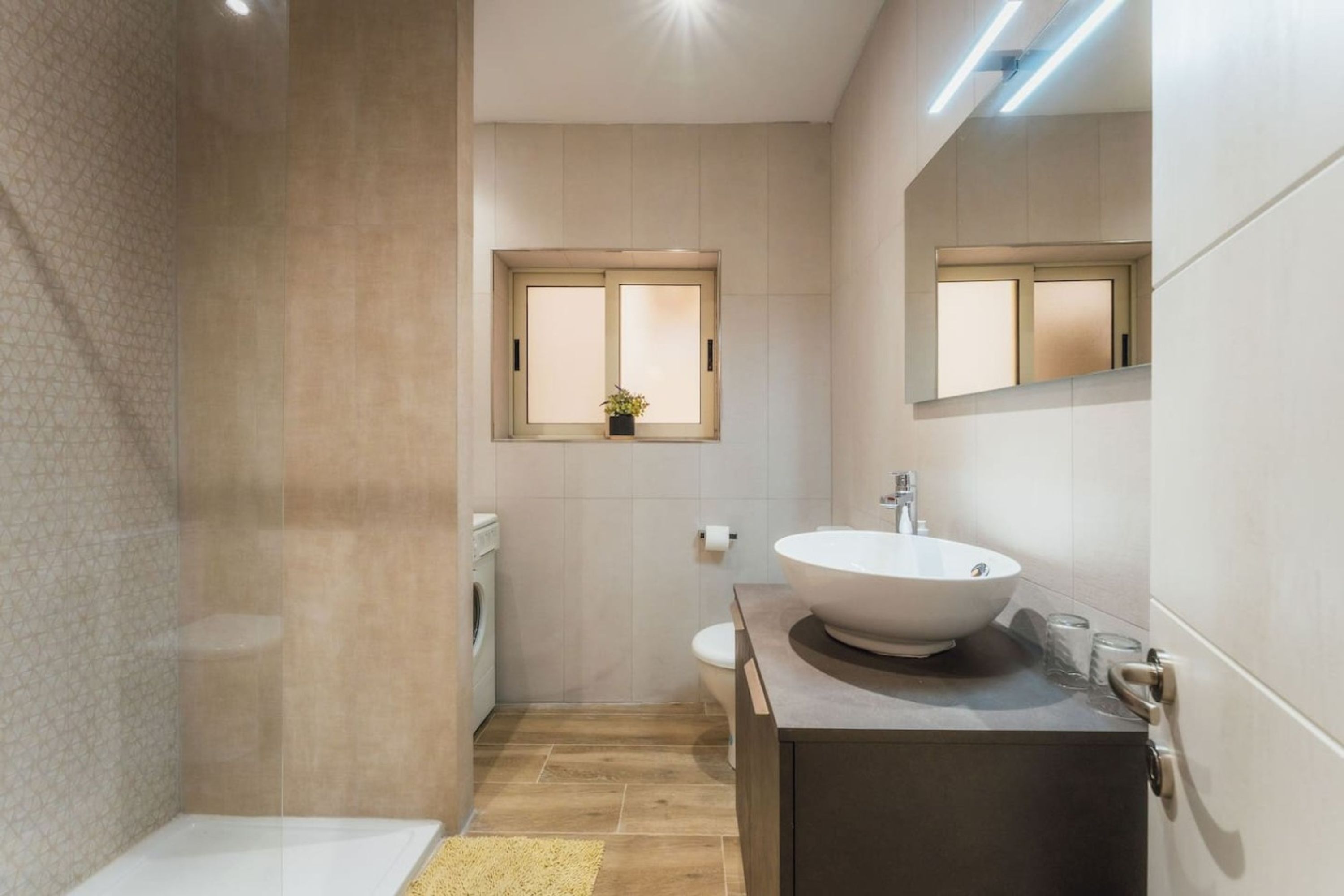 A modern bathroom with a large sink, a toilet, a washing machine, and a window with a plant.