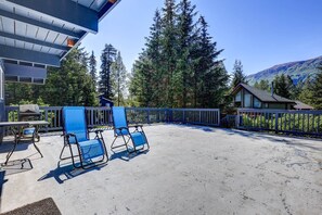 Eine Terrasse mit blauen Stühlen und einem Grill, umgeben von einer Holzterrasse und einem Metallzaun, mit Bäumen und einem Berg im Hintergrund.