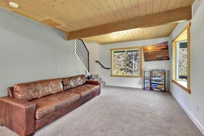 Una habitación con un sofá de cuero marrón, una escalera, un ventanal con vistas a árboles y un estante con juegos de mesa.