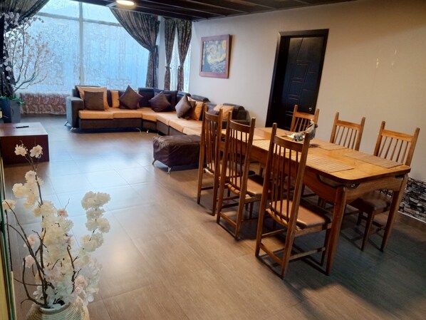 A dining area with a wooden table and chairs, a sofa with cushions, a potted plant, and a painting on the wall.