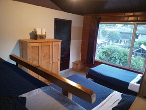 Una habitación con dos camas, un armario de madera, un mueble pequeño y una ventana que da a zonas verdes y edificios.