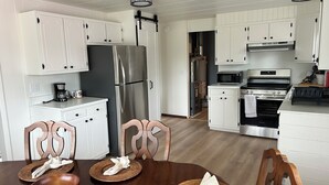 A modern kitchen with white cabinetry, stainless steel appliances, and a dining area with wooden chairs and a table set for two. - Enjoyable 3-bedroom house in charming Rogers City perfect for relaxing (Rogers City)