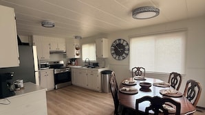 A modern kitchen with white cabinets, a dining area with a wooden table and chairs, and a large wall clock. - Enjoyable 3-bedroom house in charming Rogers City perfect for relaxing (Rogers City)