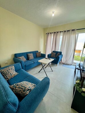 Un salón con un sofá azul, una mesita blanca y vistas a través de una ventana con cortinas.
