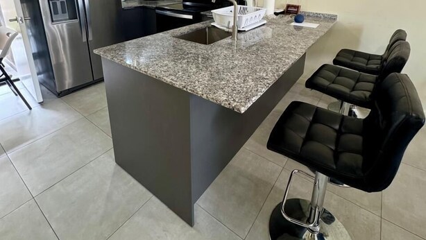 A modern kitchen with a granite countertop, stainless steel appliances, and black bar stools. - Gated Luxury Fort George Retreat | Panoramic Port-of-Spain Views | Sleeps 8+ (Port of Spain)