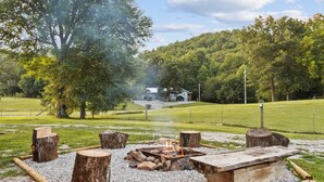 Ein Lagerfeuer mit Holzscheiten und einer Steinsitzbank in einer grünen Umgebung mit Bäumen und einem Haus im Hintergrund.