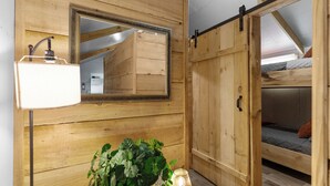 Ein Zimmer mit Holzwänden, einem Bett mit gesteppter Matratze, einem hängenden Spiegel und einer Stehlampe.