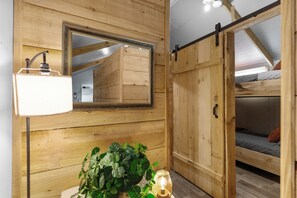 Ein Zimmer mit Holzwänden, einem Bett mit gesteppter Matratze, einem hängenden Spiegel und einer Stehlampe.