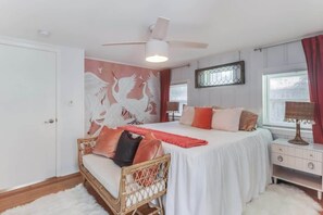 A bedroom with a bed, a ceiling fan, a wall mural, a window with curtains, and a bedside table. - The Cabana on the Coast (Gulfport)