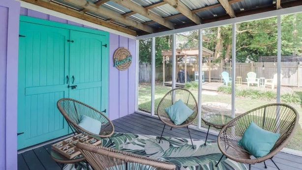 A covered patio with wicker chairs and a table, a turquoise door, and a view of the backyard. - The Little Sandy Flamingo (Gulfport)