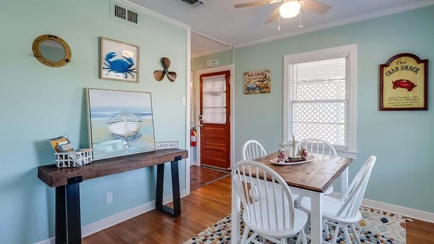A dining area with a wooden table and white chairs, a ceiling fan, and wall decorations including a framed picture of a boat and a sign that reads 'The Crab Shack.' - The Cozy Blue Cottage (Long Beach)