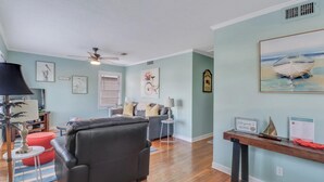 A living room with a black leather sofa, a red ottoman, a wooden coffee table, a ceiling fan, and artwork on the walls. - The Cozy Blue Cottage (Long Beach)