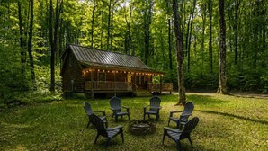 Una cabaña de troncos con un porche, rodeada por un bosque. Se observan cuatro sillas Adirondack y un brasero al frente.