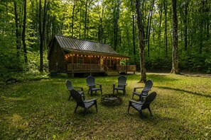 Eine Blockhütte mit einer Terrasse, umgeben von einem Wald. Im Vordergrund stehen vier Adirondack-Stühle und eine Feuerstelle.