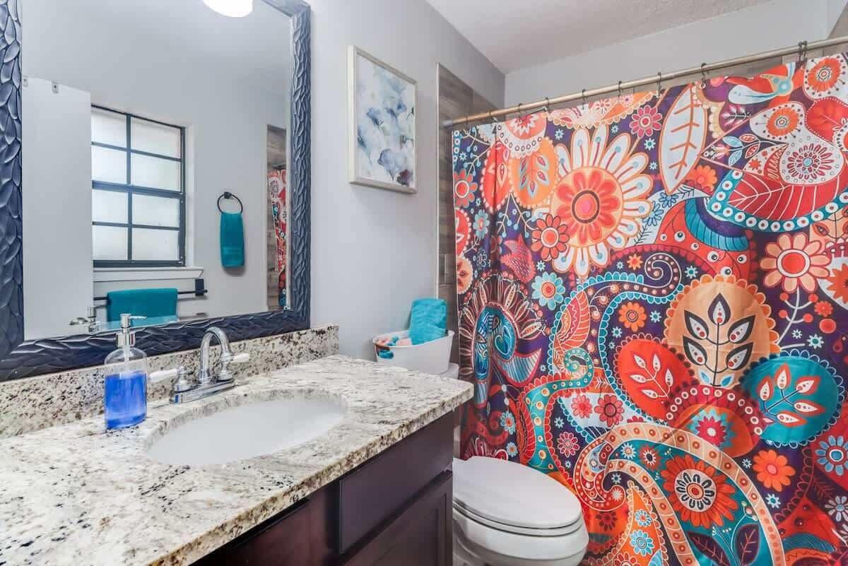 A bathroom with a large mirror, a sink with a granite countertop, a shower curtain with a colorful paisley pattern, and a white toilet.