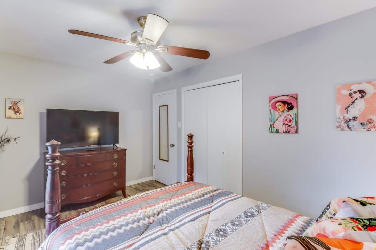 A bedroom with a bed, dresser, ceiling fan, TV, and artwork on the wall.