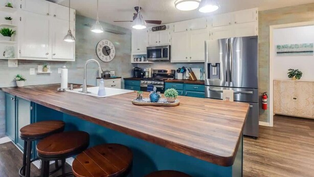 A modern kitchen with white cabinets, a central island with stools, stainless steel appliances, pendant lighting, and a clock on the wall. - The Gulf Breeze Getaway (Gulfport)