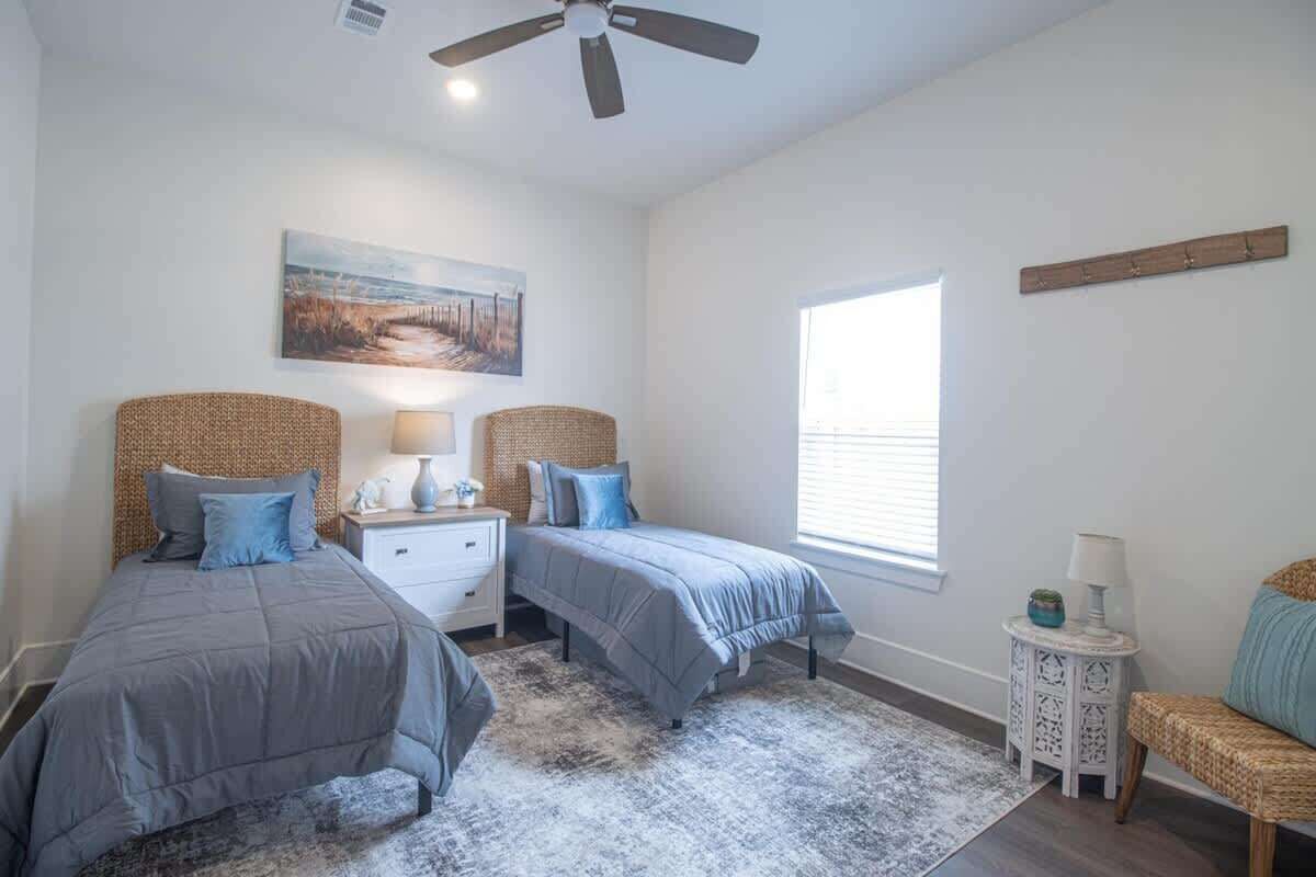 A bedroom with two beds, a ceiling fan, a painting, a window, and a small side table.