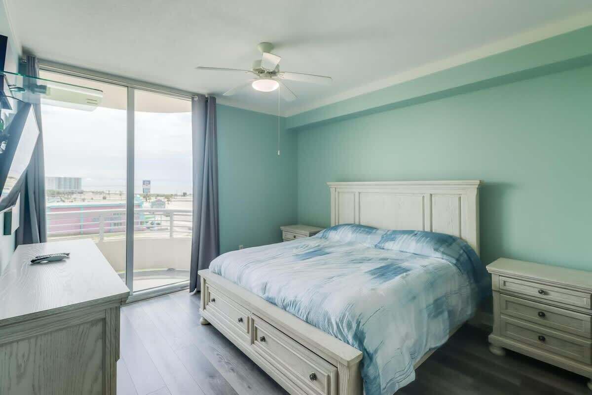 A bedroom with a bed, bedside tables, a desk, a ceiling fan, and a large window with a view.