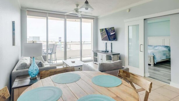 Una habitación de hotel moderna con balcón, zona de comedor con mesa y sillas de madera, televisor montado en la pared y cama con colcha azul.