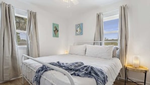 Un dormitorio con cama, cortinas, una mesita de noche con lámpara y dos cuadros en la pared.