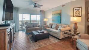 A living room with a sofa, ottoman, and a small table with a lamp. There is a ceiling fan, a TV, and a large window with curtains. - Sunsets From The Sandy Balcony (Biloxi)