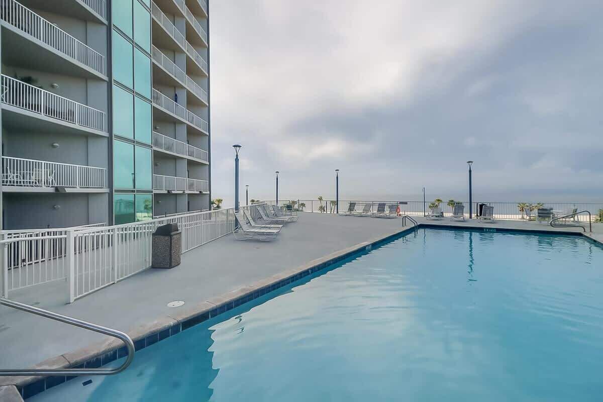 Una piscina en la azotea con sillones de descanso, un edificio con balcones y un cielo nublado.