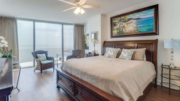 A bedroom with a large bed, a ceiling fan, a painting, a chair, a lamp, and a view of the outdoors. - Endless Blue at the Gulf Watch (Biloxi)