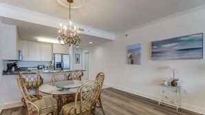 A dining area with a round table and wicker chairs, a chandelier, and a kitchen in the background. - Endless Blue at the Gulf Watch (Biloxi)