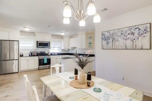 A modern kitchen with white cabinets, stainless steel appliances, and a dining area with a wooden table and chairs. - The Magnolia Retreat (Gulfport)