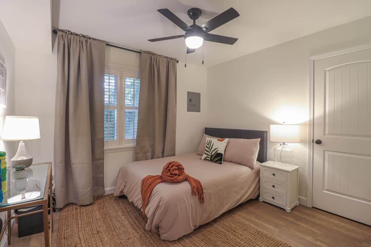 Une chambre à coucher comprenant un lit, des tables de chevet, un ventilateur de plafond, une fenêtre avec des rideaux et une porte.