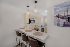 Una cocina moderna con una isla central, taburetes de bar, iluminación colgante y un cuadro de un puerto deportivo.