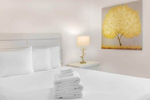 A hotel room with a neatly made bed, a bedside lamp, and a painting of a tree. - Cozy 2 Bed/1.5 Bath Duplex Just Blocks From Downtown Biloxi! (Biloxi)