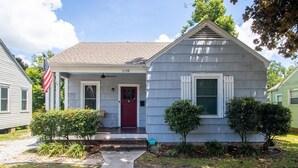A single-story house with a front porch, a red door, and a small front yard with two bushes. - Cottage on Broadmoor (Gulfport)