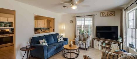 Un salon comprenant un canapé, un fauteuil, un téléviseur, un ventilateur de plafond et un coin cuisine.