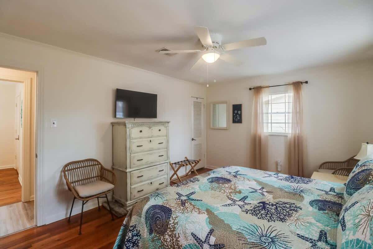 Ein Schlafzimmer mit Bett, Deckenventilator, Kommode, Stuhl, Fernseher und einem Fenster mit Vorhängen.