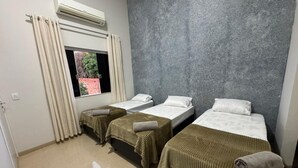 A hotel room with three beds, a window with curtains, an air conditioning unit, and a grey textured wall. - Alvorada Hospedagem (Jataí)