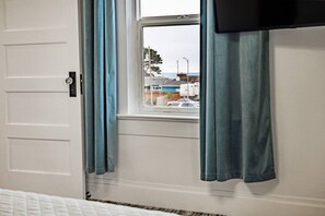 Ein Zimmer mit weißer Tür, einem Fenster mit blauen Vorhängen und Blick auf eine Straße mit Gebäuden und einem Auto.