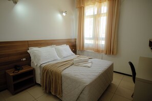 Una cama bien hecha con cabecera de madera, una mesita de noche con un teléfono y una ventana con cortinas traslúcidas.