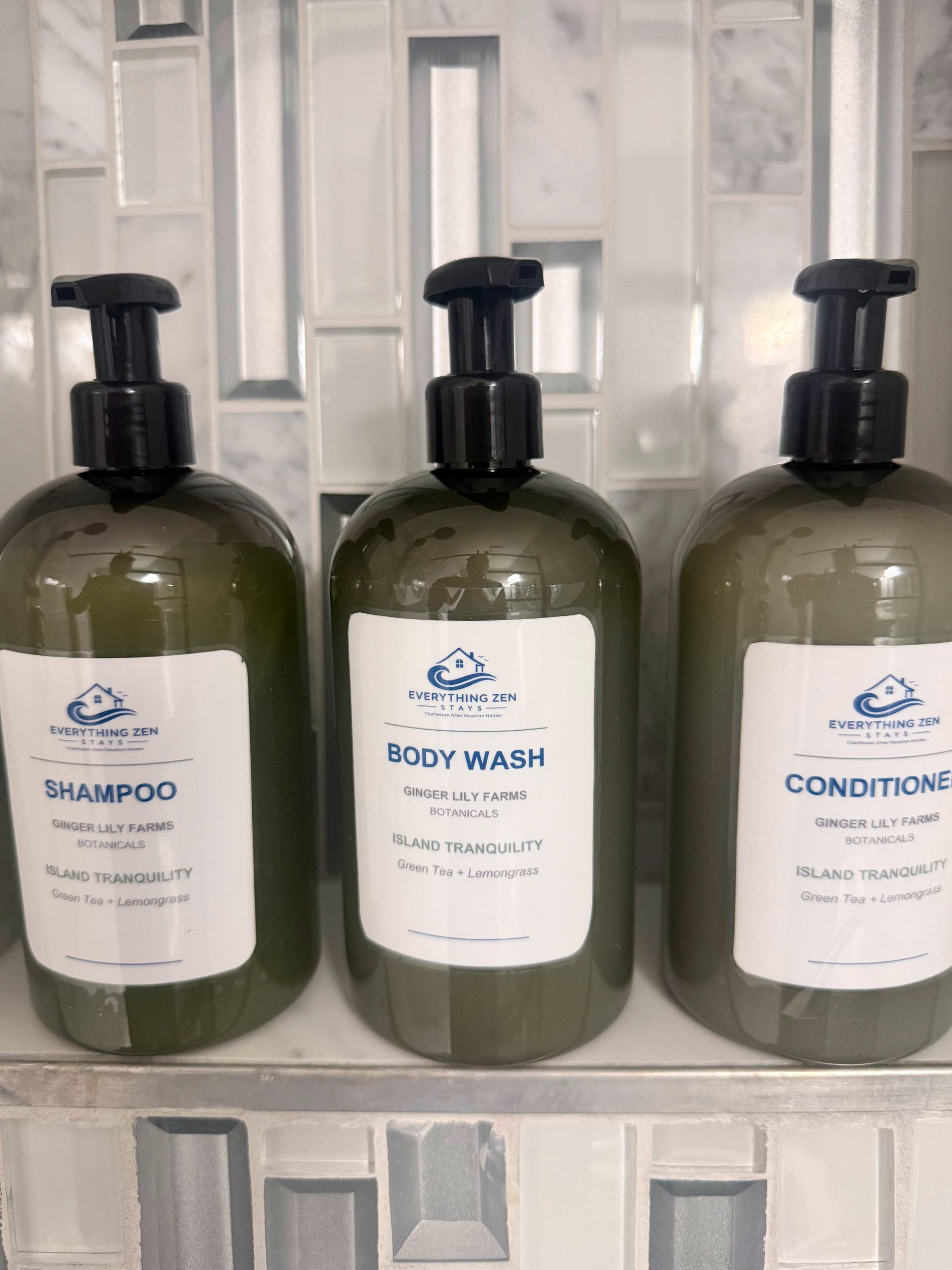 Drei Flaschen Duschgel und Shampoo mit Etiketten auf einer gefliesten Oberfläche.