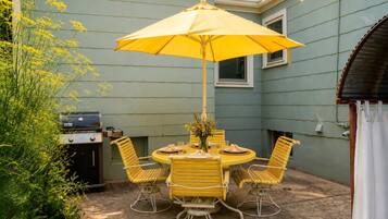 An outdoor patio with a round table, yellow chairs, a yellow umbrella, and a grill.