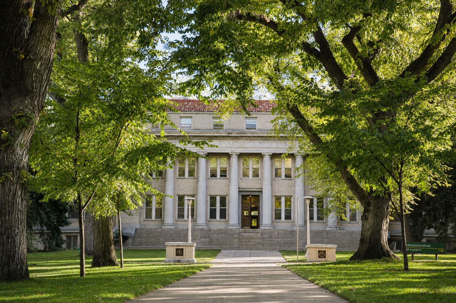 A stately building with a central entrance, flanked by large trees, and a paved pathway leading to the entrance.