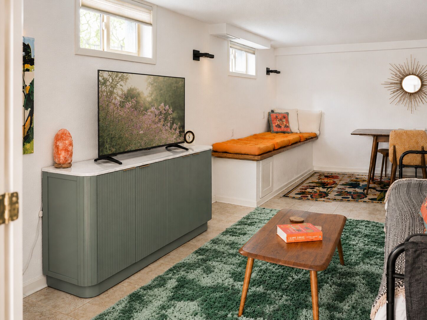 Un espace de vie compact comprenant un téléviseur posé sur un meuble, un petit coin salon avec des coussins, une table à manger et un tapis vert.