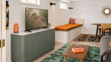 Un espace de vie compact comprenant un téléviseur posé sur un meuble, un petit coin salon avec des coussins, une table à manger et un tapis vert.