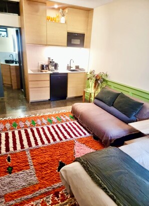 Una habitación moderna con una cama, un sofá, una mesita con un florero y una kitchenette al fondo.
