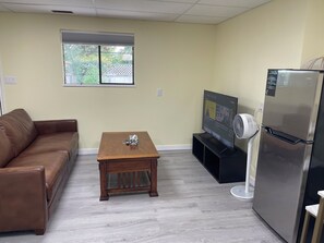 Ein Wohnzimmer mit braunem Ledersofa, einem Holztisch, einem Flachbildfernseher, einem weißen Standventilator und einem Edelstahlkühlschrank.