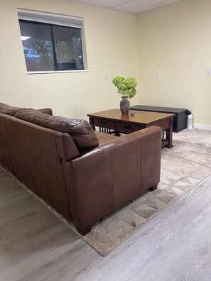 A living room with a brown leather sofa, a wooden coffee table, a vase with green plants, and a window with blinds. - Anna's cozy house (Delta)