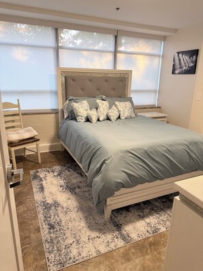 Ein Schlafzimmer mit Bett, Stuhl, Nachttisch, Schreibtisch, Teppich und einem Fenster mit Jalousien.